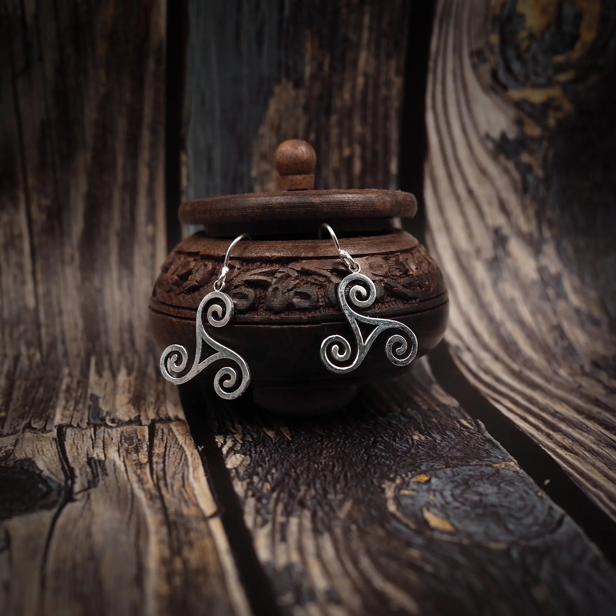 In the picture you can see a pair of silver coloured earrings, shaped like Triglavs, also called triunity symbols. The earrings are hanging from a small, round, decorated wooden box.