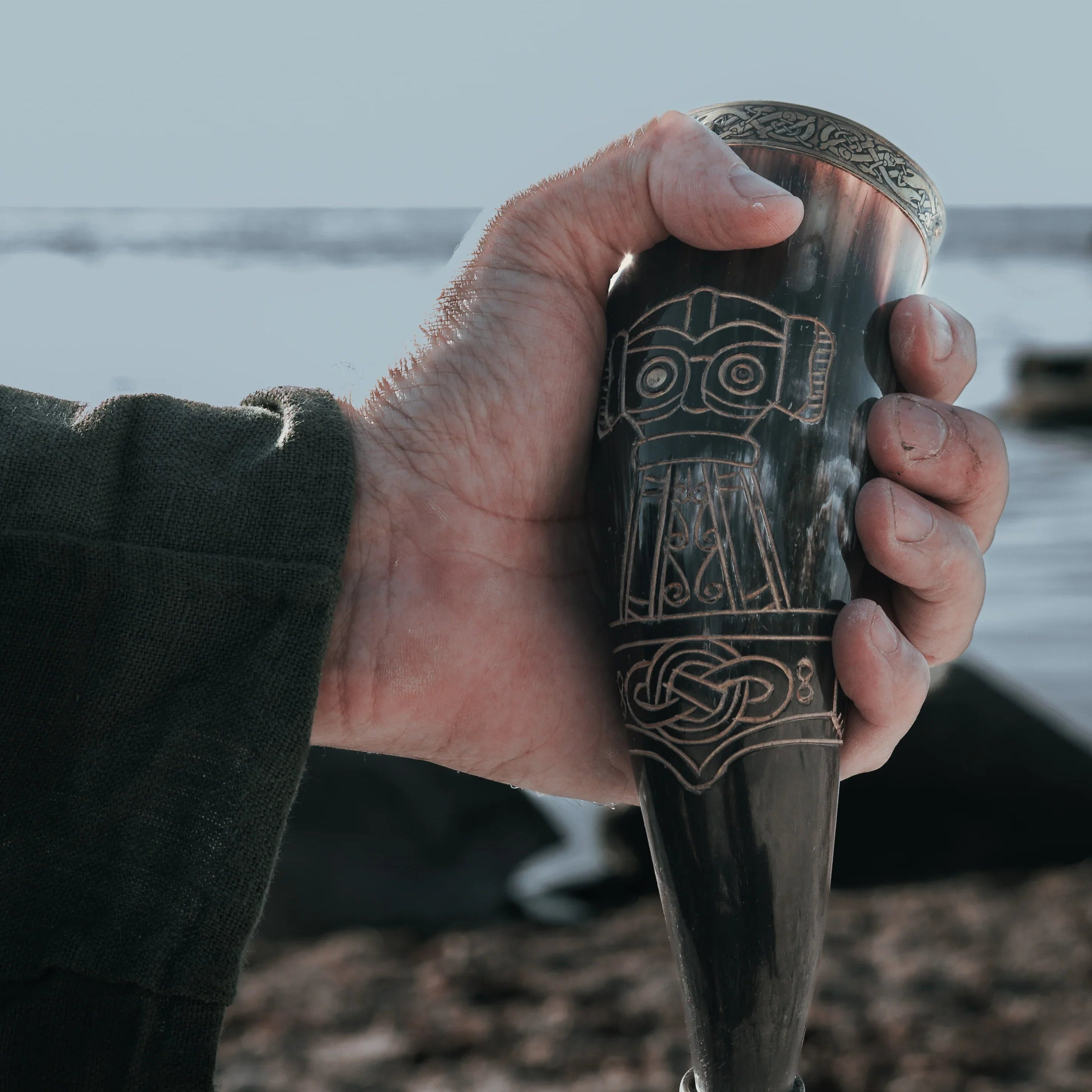 Dryckeshorn med korp och gravyr gjord av organiskt horn, cirka 250 ml, med mässingband och Viking-inspireade mönster, hållen i hand mot hav och klippor i bakgrunden