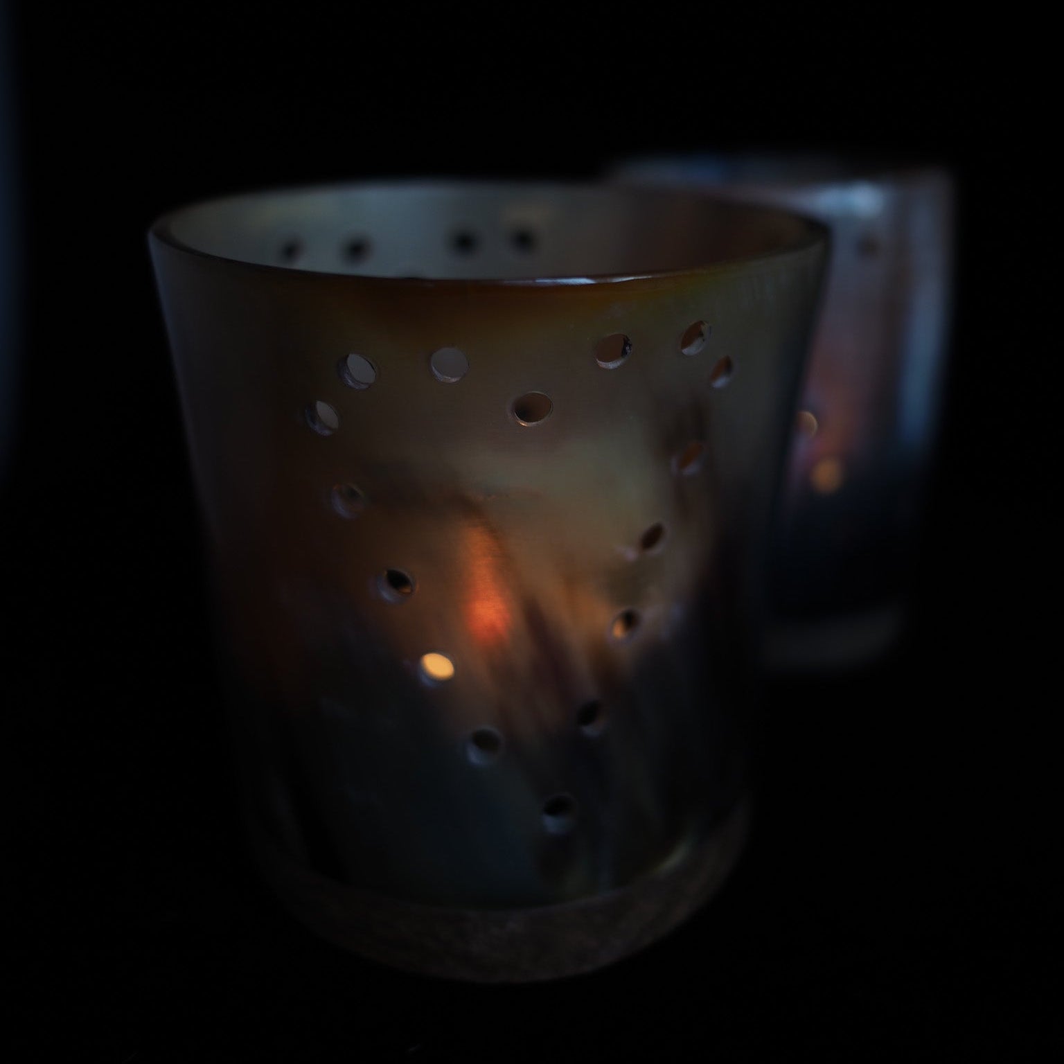 Mörkfärgad bockhornsljuslykta med cirkulära hål, upplysta från insidan av ett ljus som skapar en varm orangegul glöd. Två horn ljuslyktor för ljusslinga.