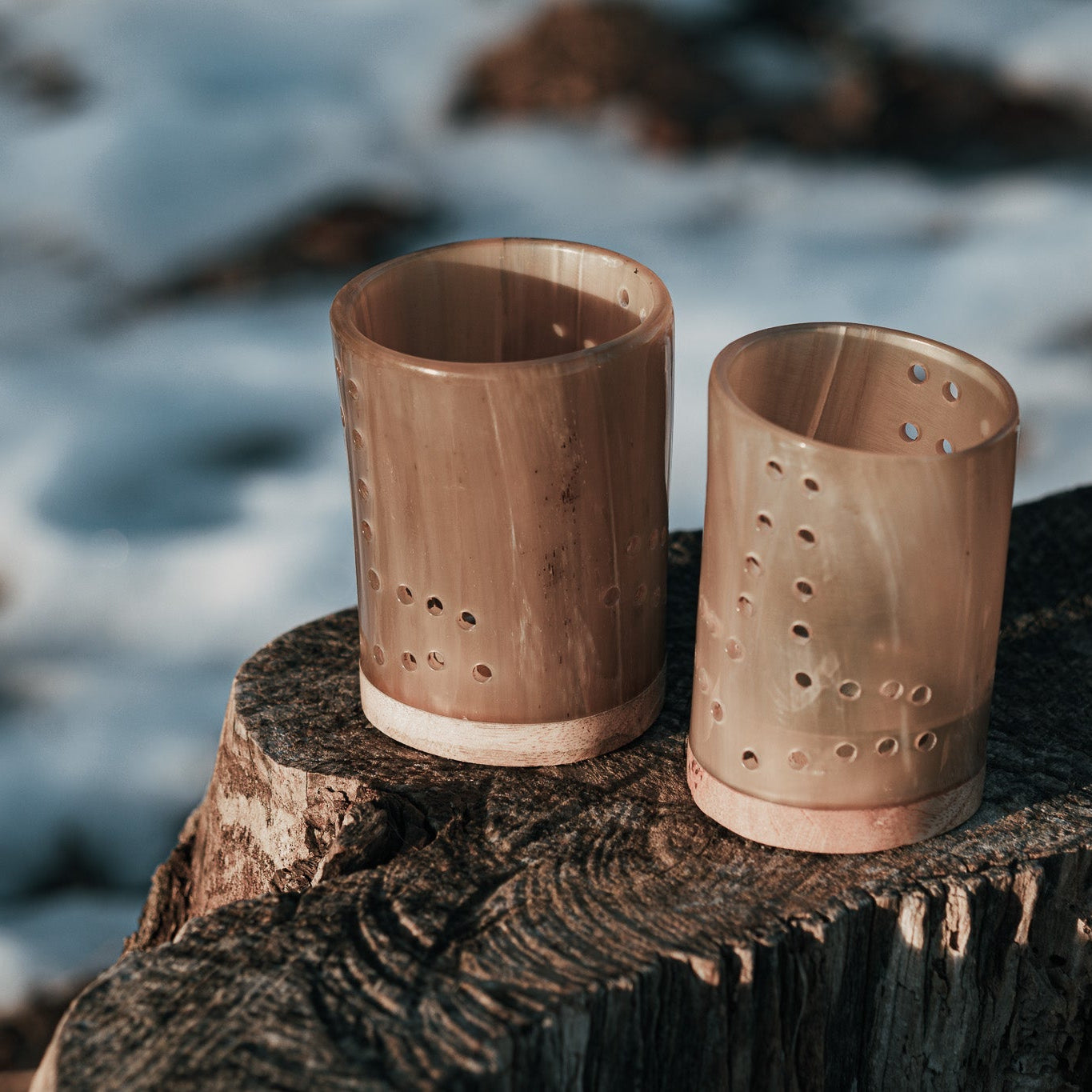 Två cylindriska ljuslyktor av organiskt horn med dekorativa hål i hjärtmönster, ställda på en vädersliten trästock framför havet. Naturlig hornfärg med varmt brunt patina och vita läderband i basen.